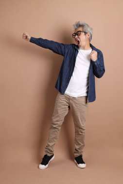 Gözlüklü, Asyalı, enerjik, üst düzey bir adam kahverengi arka planda izole edilmiş ve günlük kıyafetlerle poz veriyor. Olgun adam, Asyalı adam. Yaşlı, stüdyo fotoğrafı. Çoklu duygular.