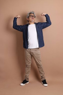 Gözlüklü, Asyalı, enerjik, üst düzey bir adam kahverengi arka planda izole edilmiş ve günlük kıyafetlerle poz veriyor. Olgun adam, Asyalı adam. Yaşlı, stüdyo fotoğrafı. Çoklu duygular.