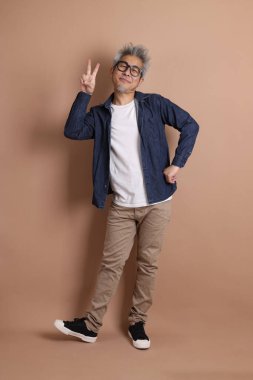 Gözlüklü, Asyalı, enerjik, üst düzey bir adam kahverengi arka planda izole edilmiş ve günlük kıyafetlerle poz veriyor. Olgun adam, Asyalı adam. Yaşlı, stüdyo fotoğrafı. Çoklu duygular.