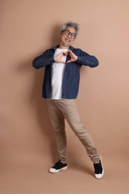 Gözlüklü, Asyalı, enerjik, üst düzey bir adam kahverengi arka planda izole edilmiş ve günlük kıyafetlerle poz veriyor. Olgun adam, Asyalı adam. Yaşlı, stüdyo fotoğrafı. Çoklu duygular.