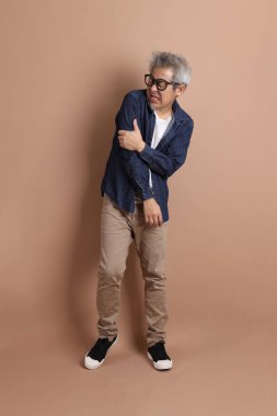 Gözlüklü, Asyalı, enerjik, üst düzey bir adam kahverengi arka planda izole edilmiş ve günlük kıyafetlerle poz veriyor. Olgun adam, Asyalı adam. Yaşlı, stüdyo fotoğrafı. Çoklu duygular.