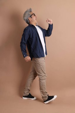 Gözlüklü, Asyalı, enerjik, üst düzey bir adam kahverengi arka planda izole edilmiş ve günlük kıyafetlerle poz veriyor. Olgun adam, Asyalı adam. Yaşlı, stüdyo fotoğrafı. Çoklu duygular.