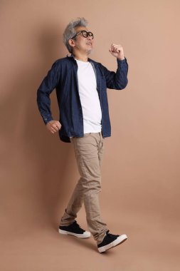 Gözlüklü, Asyalı, enerjik, üst düzey bir adam kahverengi arka planda izole edilmiş ve günlük kıyafetlerle poz veriyor. Olgun adam, Asyalı adam. Yaşlı, stüdyo fotoğrafı. Çoklu duygular.