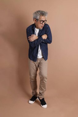 Gözlüklü, Asyalı, enerjik, üst düzey bir adam kahverengi arka planda izole edilmiş ve günlük kıyafetlerle poz veriyor. Olgun adam, Asyalı adam. Yaşlı, stüdyo fotoğrafı. Çoklu duygular.