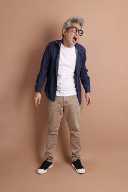 Gözlüklü, Asyalı, enerjik, üst düzey bir adam kahverengi arka planda izole edilmiş ve günlük kıyafetlerle poz veriyor. Olgun adam, Asyalı adam. Yaşlı, stüdyo fotoğrafı. Çoklu duygular.