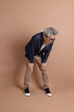 Gözlüklü, Asyalı, enerjik, üst düzey bir adam kahverengi arka planda izole edilmiş ve günlük kıyafetlerle poz veriyor. Olgun adam, Asyalı adam. Yaşlı, stüdyo fotoğrafı. Çoklu duygular.