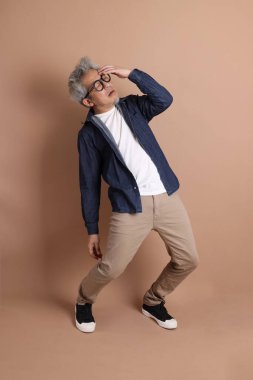 Gözlüklü, Asyalı, enerjik, üst düzey bir adam kahverengi arka planda izole edilmiş ve günlük kıyafetlerle poz veriyor. Olgun adam, Asyalı adam. Yaşlı, stüdyo fotoğrafı. Çoklu duygular.