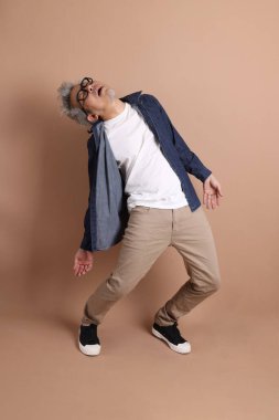 Gözlüklü, Asyalı, enerjik, üst düzey bir adam kahverengi arka planda izole edilmiş ve günlük kıyafetlerle poz veriyor. Olgun adam, Asyalı adam. Yaşlı, stüdyo fotoğrafı. Çoklu duygular.