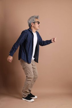 Gözlüklü, Asyalı, enerjik, üst düzey bir adam kahverengi arka planda izole edilmiş ve günlük kıyafetlerle poz veriyor. Olgun adam, Asyalı adam. Yaşlı, stüdyo fotoğrafı. Çoklu duygular.