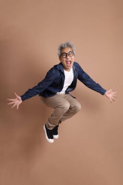 Gözlüklü, Asyalı, enerjik, üst düzey bir adam kahverengi arka planda izole edilmiş ve günlük kıyafetlerle poz veriyor. Olgun adam, Asyalı adam. Yaşlı, stüdyo fotoğrafı. Çoklu duygular.