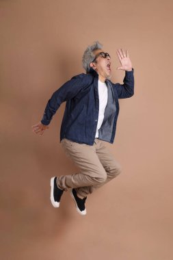 Gözlüklü, Asyalı, enerjik, üst düzey bir adam kahverengi arka planda izole edilmiş ve günlük kıyafetlerle poz veriyor. Olgun adam, Asyalı adam. Yaşlı, stüdyo fotoğrafı. Çoklu duygular.