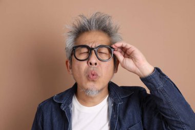 Gözlüklü, Asyalı, enerjik, üst düzey bir adam kahverengi arka planda izole edilmiş ve günlük kıyafetlerle poz veriyor. Olgun adam, Asyalı adam. Yaşlı, stüdyo fotoğrafı. Çoklu duygular