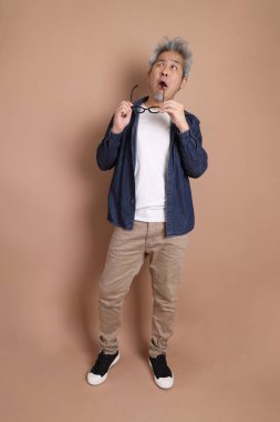 Gözlüklü, Asyalı, enerjik, üst düzey bir adam kahverengi arka planda izole edilmiş ve günlük kıyafetlerle poz veriyor. Olgun adam, Asyalı adam. Yaşlı, stüdyo fotoğrafı. Çoklu duygular