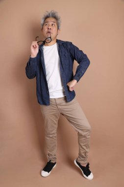 Gözlüklü, Asyalı, enerjik, üst düzey bir adam kahverengi arka planda izole edilmiş ve günlük kıyafetlerle poz veriyor. Olgun adam, Asyalı adam. Yaşlı, stüdyo fotoğrafı. Çoklu duygular