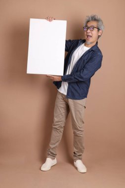 Elinde boş bir tuval tutan, gözlüklü, Asyalı, enerjik, üst düzey bir adam. Kahverengi arka planda, günlük kıyafetlerle izole edilmiş. Olgun adam, Asyalı adam. Yaşlı, stüdyo fotoğrafı. Çoklu duygular. 