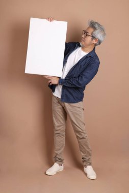 Elinde boş bir tuval tutan, gözlüklü, Asyalı, enerjik, üst düzey bir adam. Kahverengi arka planda, günlük kıyafetlerle izole edilmiş. Olgun adam, Asyalı adam. Yaşlı, stüdyo fotoğrafı. Çoklu duygular. 
