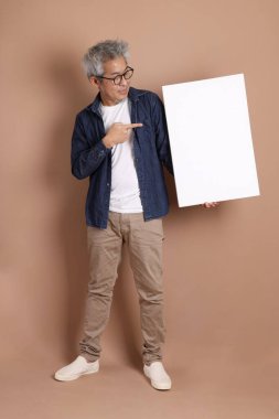 Elinde boş bir tuval tutan, gözlüklü, Asyalı, enerjik, üst düzey bir adam. Kahverengi arka planda, günlük kıyafetlerle izole edilmiş. Olgun adam, Asyalı adam. Yaşlı, stüdyo fotoğrafı. Çoklu duygular. 
