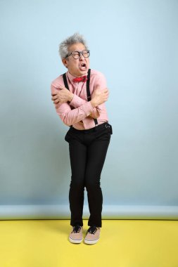 Asyalı, siyah pantolon askısı takan, kırmızı fiyonklu, mavi arka planda soğuk hissetme hareketi yapan son sınıf öğrencisi. Sevgililer Günü