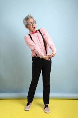 Asyalı, siyah pantolon askısı takmış, kırmızı pruvalı, yorgun ve tembel hareketleriyle mavi arka planda izole edilmiş. Sevgililer Günü