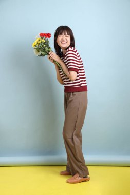 Mavi arka planda bir buket çiçek saklama jesti ile Breton gömlekli neşeli, genç Asyalı kadın. Sevgililer Günü, Kadınlar Günü, Doğum Günü