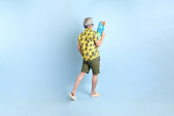 С Днем Сон-Крэйн. Азиатский энергичный пожилой человек в летней одежде и солнцезащитных очках с жестом держания пластикового водяного пистолета изолирован на синем фоне. Фестиваль Сонгкран. Новый год в Таиланде.