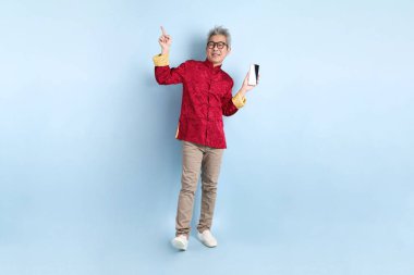 Yeni Çin yılın kutlu olsun. Asya 'nın enerjik kıdemli adamı kırmızı geleneksel Cheongsam Qipao ya da Changshan elbisesi giyiyor ve mavi arka planda cep telefonlarını izole ediyor..