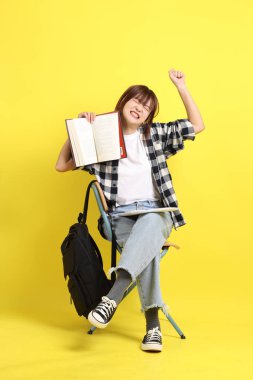 Ekose gömlek giyen Asyalı kız öğrenci okul çantası ve kitap taşıyor, oturma ve çalışma jesti. Yurtdışında okumak, Z Geni, E-öğrenimi.