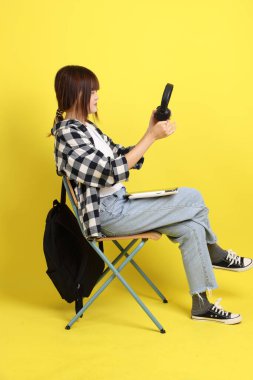 Ekose gömlek giyen Asyalı kız öğrenci okul çantası ve kitap taşıyor, oturma ve kulaklık takma hareketi yapıyor. Yurtdışında okumak, Z Geni, E-öğrenimi.