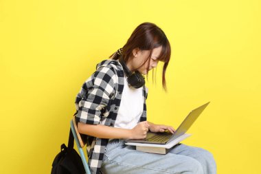 Ekose gömlek giyen Asyalı kız öğrenci okul çantası ve kitap taşıyor. Oturma eylemi ve dizüstü bilgisayar tutuyor. Yurtdışında okumak, Z Geni, E-öğrenimi.