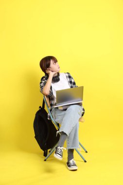 Ekose gömlek giyen Asyalı kız öğrenci okul çantası ve kitap taşıyor. Oturma eylemi ve dizüstü bilgisayar tutuyor. Yurtdışında okumak, Z Geni, E-öğrenimi.