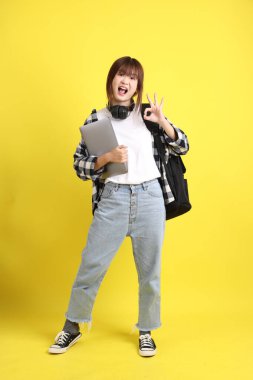 Ekose gömlek giyen Asyalı kız öğrenci okul çantası ve kitap taşıyor, ayakta durma hareketi ve dizüstü bilgisayar tutuyor. Yurtdışında okumak, Z Geni, E-öğrenimi.