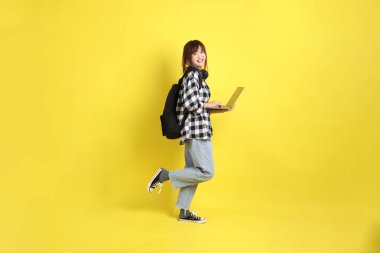 Ekose gömlek giyen Asyalı kız öğrenci okul çantası ve kitap taşıyor, ayakta durma hareketi ve dizüstü bilgisayar tutuyor. Yurtdışında okumak, Z Geni, E-öğrenimi.