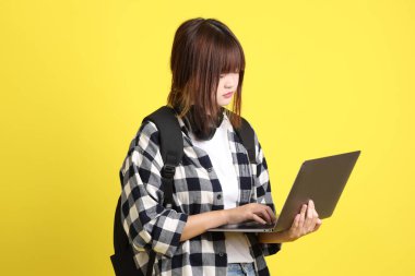 Ekose gömlek giyen Asyalı kız öğrenci okul çantası ve kitap taşıyor, ayakta durma hareketi ve dizüstü bilgisayar tutuyor. Yurtdışında okumak, Z Geni, E-öğrenimi.