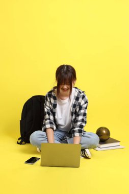 Ekose gömlek giyen Asyalı kız öğrenci okul çantası ve kitap taşıyor. Oturma ve dizüstü bilgisayarla çalışma jesti. Yurtdışında okumak, Z Geni, E-öğrenimi.