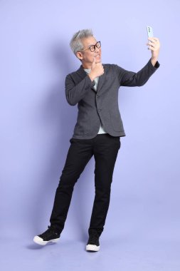 50 'li yılların Asyalı kıdemli iş adamı. Gözlüklü, resmi giyinmiş. Mor arka planda izole edilmiş cep telefonu bulundurma jesti.. 