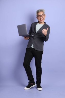 50 'li yılların Asyalı kıdemli iş adamı, gözlüklü, resmi giyinmiş ve dizüstü bilgisayarını mor arka planda tutuyormuş.. 