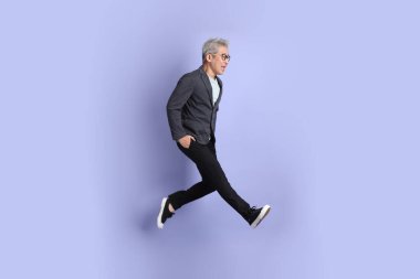 50 'lerin Asyalı kıdemli iş adamı, gözlüklü, resmi giyinmiş, mor arka planda izole bir şekilde atlama hareketi yapıyor.. 