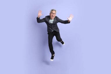 50 'lerin Asyalı kıdemli iş adamı, gözlüklü, resmi giyinmiş, mor arka planda izole bir şekilde atlama hareketi yapıyor.. 