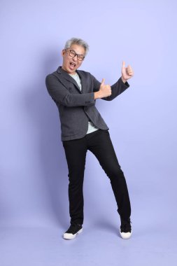 50 'li yılların Asyalı kıdemli iş adamı, gözlüklü, resmi kıyafetli, mor arka planda izole edilmiş baş parmak hareketleriyle.. 