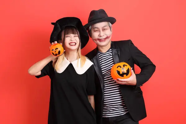 Cadılar Bayramı kostümlü Asyalı cadı kadın ve adam gülümseyen bir ifadeyle el ele tutuşurken turuncu arka planda balkabağı izole edilmiş..