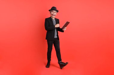 Cadılar Bayramı kostümlü Asyalı son sınıf öğrencisi. Turuncu arka planda izole edilmiş el ele tutuşan tablet jesti..