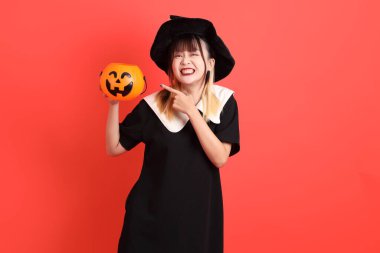 Cadılar Bayramı kostümlü genç Asyalı kadın turuncu arka planda izole edilmiş balkabağını tutan eli işaret ediyor. Ekim. 