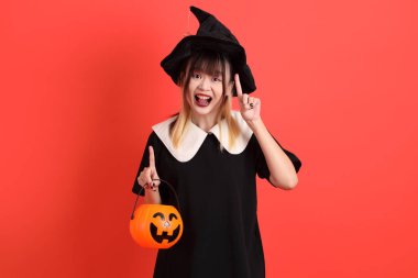 Cadılar Bayramı kostümlü genç Asyalı kadın turuncu arka planda izole edilmiş balkabağını tutan eli işaret ediyor. Ekim. 