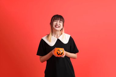 Cadılar Bayramı kostümlü genç Asyalı kadın gülümseyip ayakta dururken turuncu arka planda balkabağı tutuyor. Ekim. 