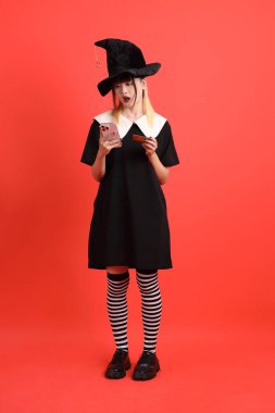 Cadıların Cadılar Bayramı kostümlü genç Asyalı kadın. Portakal rengi arka planda cep telefonu olan Holding kredi kartı jesti. Ekim