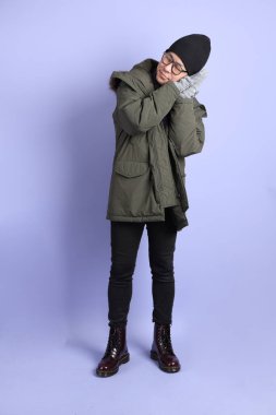 Asyalı son sınıf öğrencisi kışlık kıyafetler giyer ve mor arka planda uykulu bir jest yapar..