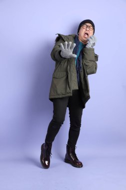 Asyalı son sınıf öğrencisi, mor arka planda izole edilmiş korku jestleriyle kışlık kıyafetler giyer..