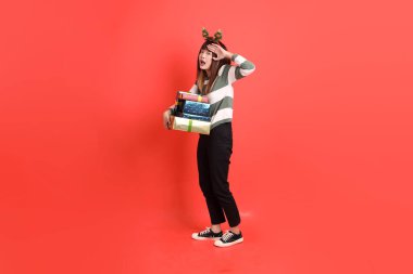 Noel Baba şapkalı ya da kışlık kıyafetler giyen Asyalı bir kadın, elinde hediyeler tutarak uzaktan bakma jestiyle, kırmızı arka planda izole bir şekilde uzaklara bakıyor. Noel Baba! Noel Baba!.