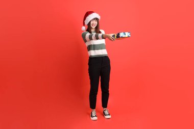 Noel Baba şapkalı ya da kışlık kıyafet giyen Asyalı bir kadın, hediye verme jestiyle elinde bir yığın hediyeyle kırmızı arka planda izole edilmiş halde bulunuyor. Noel Baba! Noel Baba!.