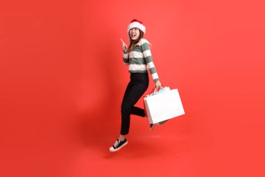 Kırmızı arka planda Noel Baba şapkalı ve alışveriş çantası taşıyan Asyalı bir kadın. Noel Baba! Noel Baba!.